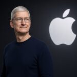 Tim Cook last day as Apple CEO: portrait of Tim Cook in a navy sweater standing beside a glowing white Apple logo on a dark stage background.