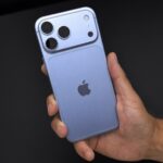 Glacier blue iPhone 18 Pro Max held in a right hand against a black desk, showing the rear camera bar and Apple logo.