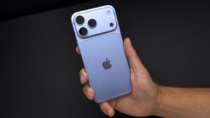 Glacier blue iPhone 18 Pro Max held in a right hand against a black desk, showing the rear camera bar and Apple logo.