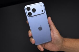 Glacier blue iPhone 18 Pro Max held in a right hand against a black desk, showing the rear camera bar and Apple logo.