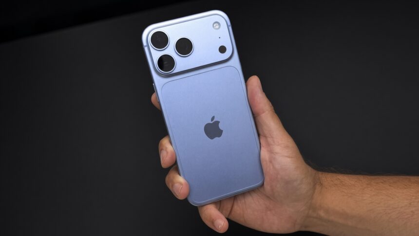 Glacier blue iPhone 18 Pro Max held in a right hand against a black desk, showing the rear camera bar and Apple logo.